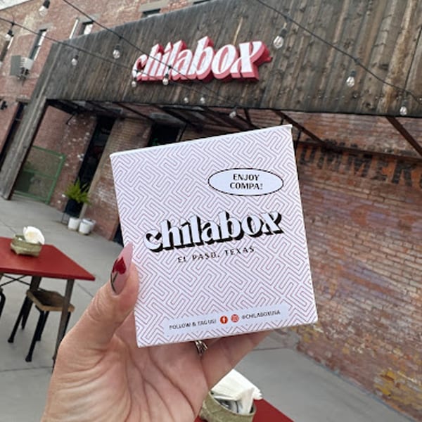 Hand holding a Chilabox branded box with the restaurant's sign and outdoor seating in the background.