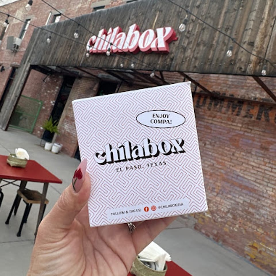 Hand holding a Chilabox branded box with the restaurant's sign and outdoor seating in the background.