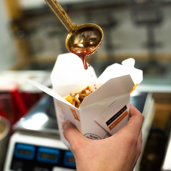 A ladle pours red sauce over Mexican food in a white takeout box, held by a hand.