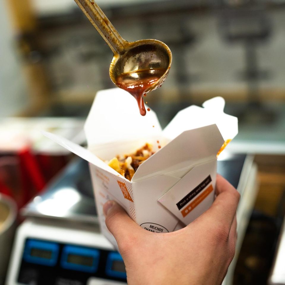 A ladle pours red sauce over Mexican food in a white takeout box, held by a hand.
